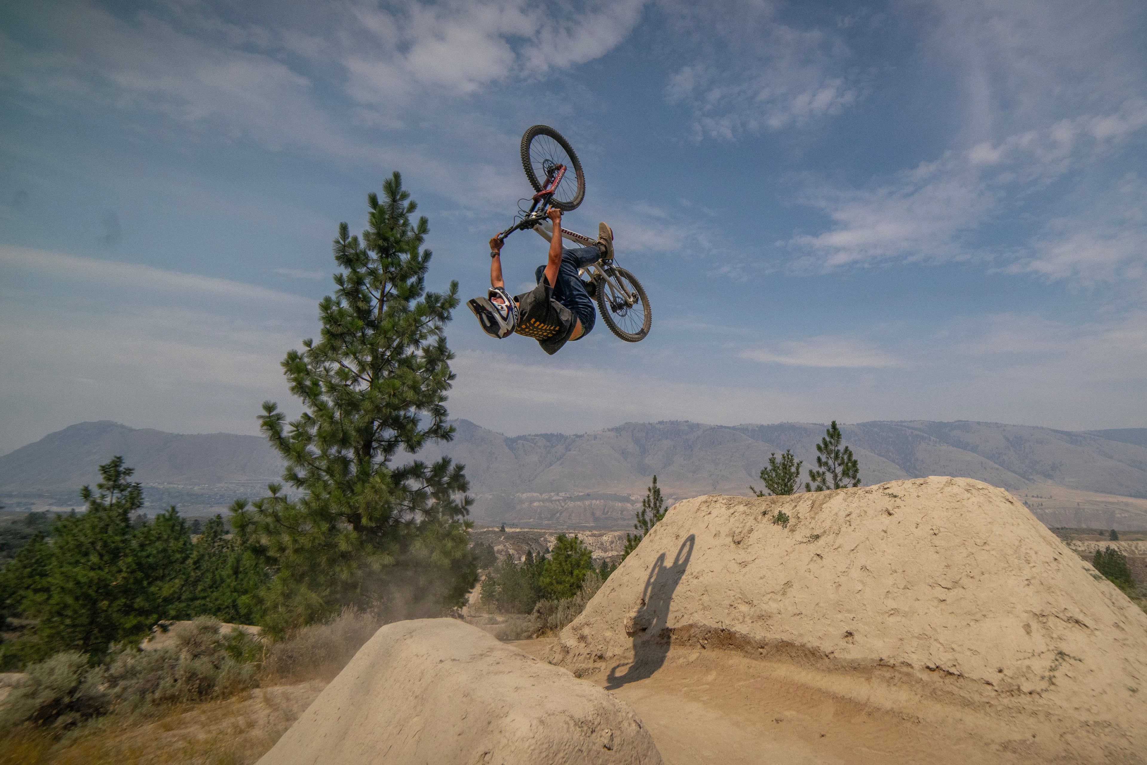 A mountain biker mid-air performing a trick on a dirt jump track, with mountains, trees, and a blue sky in the background.