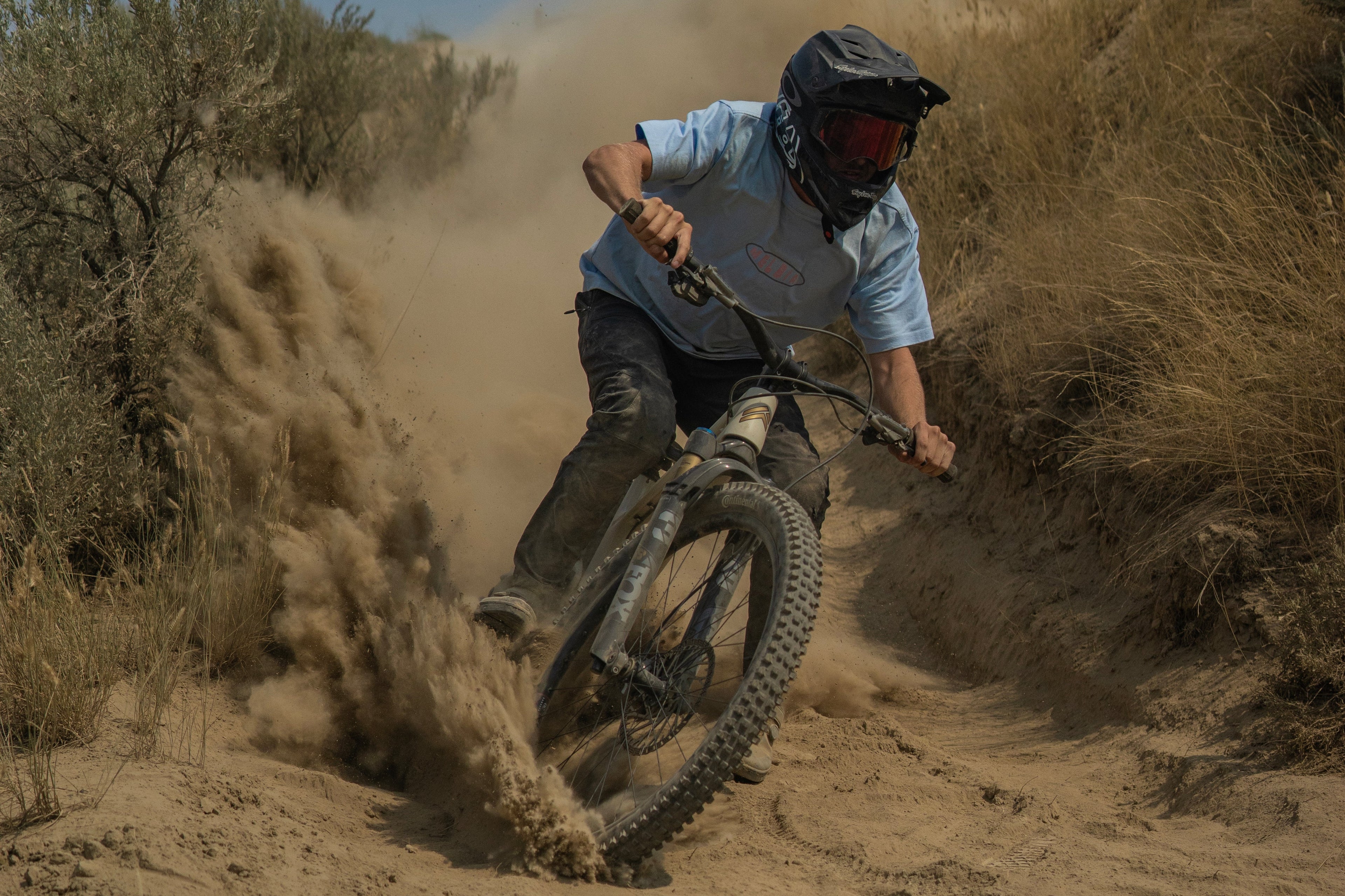 A mountain biker wearing a light blue shirt, black pants, and a helmet rides a dirt bike through a dusty sandy trail, kicking up a large cloud of sand in an outdoor desert environment.