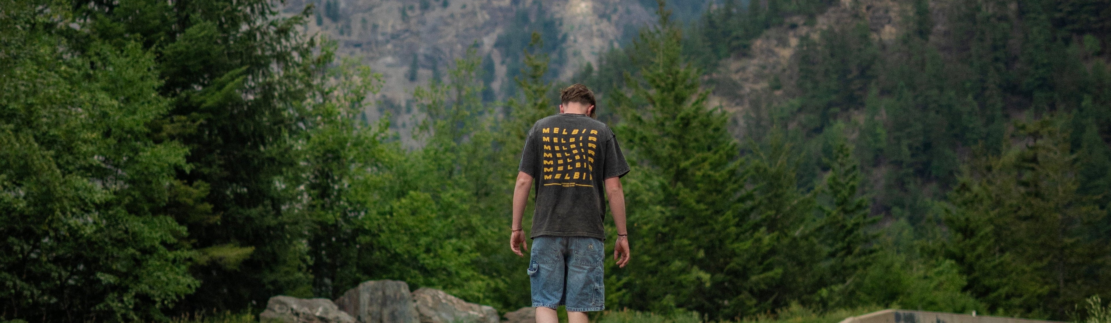 A man walks away on a paved road between large green mountains under a cloudy sky, with yellow double lines in the foreground.